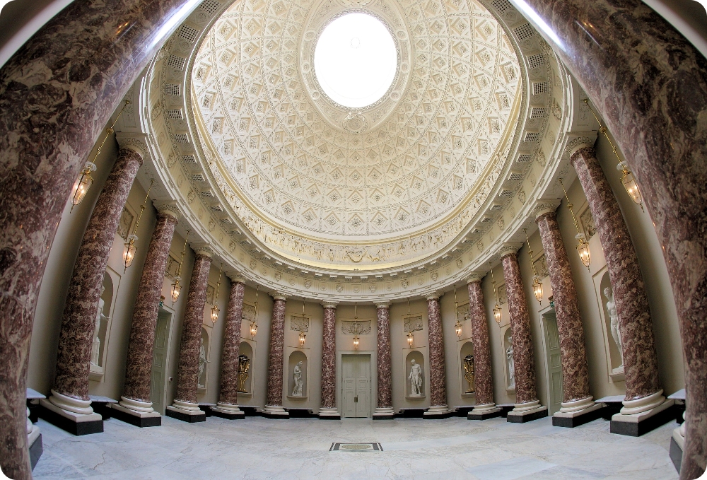 A wide oval shaped room has large columns all around in red marble effect. A huge domed rood with intricate coffers rises to a glass oculus. There are niches between the columns which contain life size large statues of roman goddesses or important political figures. There is a white marble floor.