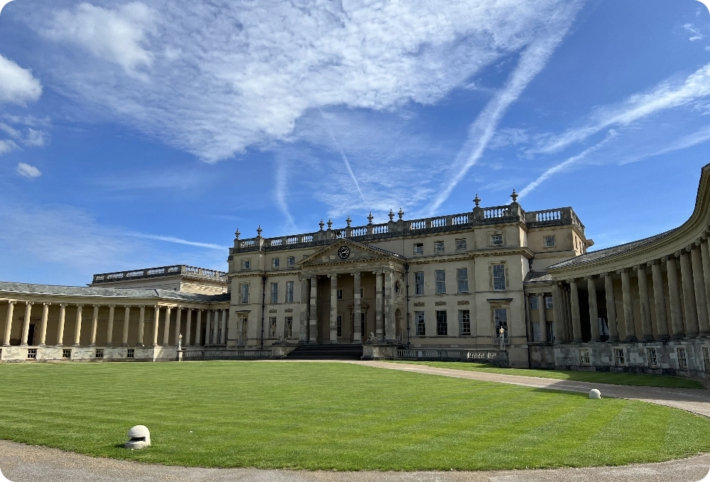 A large Palladian mansion of golden coloured stone. Centre is a rectangular building, with three storeys of windows, atop with balustrades. a porch protrudes with a clock and large columns with steps leading down to the ground. Either side of the mansion sweeps two curved wings with walkways covered all with many columns. the grass is green on the lawn and the sky is blue for the summer.
