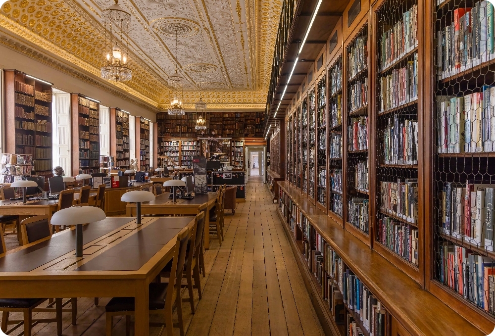 A large long rectangular library with mahogany bookcases all along the right hand side wall and south wall. Aview through the door on the other side of the room shows a connecting view through all of the state rooms. in the centre of the rooms are tables and chairs with lamps for studying/ The ceiling is covered in ornate plasterwork and has gold gilding. Three hanging glass lanterns shine.