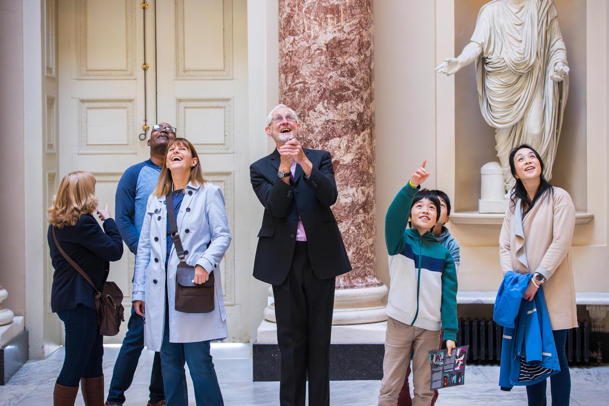 A gentleman tour guide in his 70s, tall, wearing a black suite and pink shirt, glasses with white hair and beard smiles as he points a laser to the ceiling of the Marble Saloon to show visitors a hidden item. The all look up and are smiling and laughing. There are two middle aged ladies, and a gentleman alongside a family of a woman and two young boys