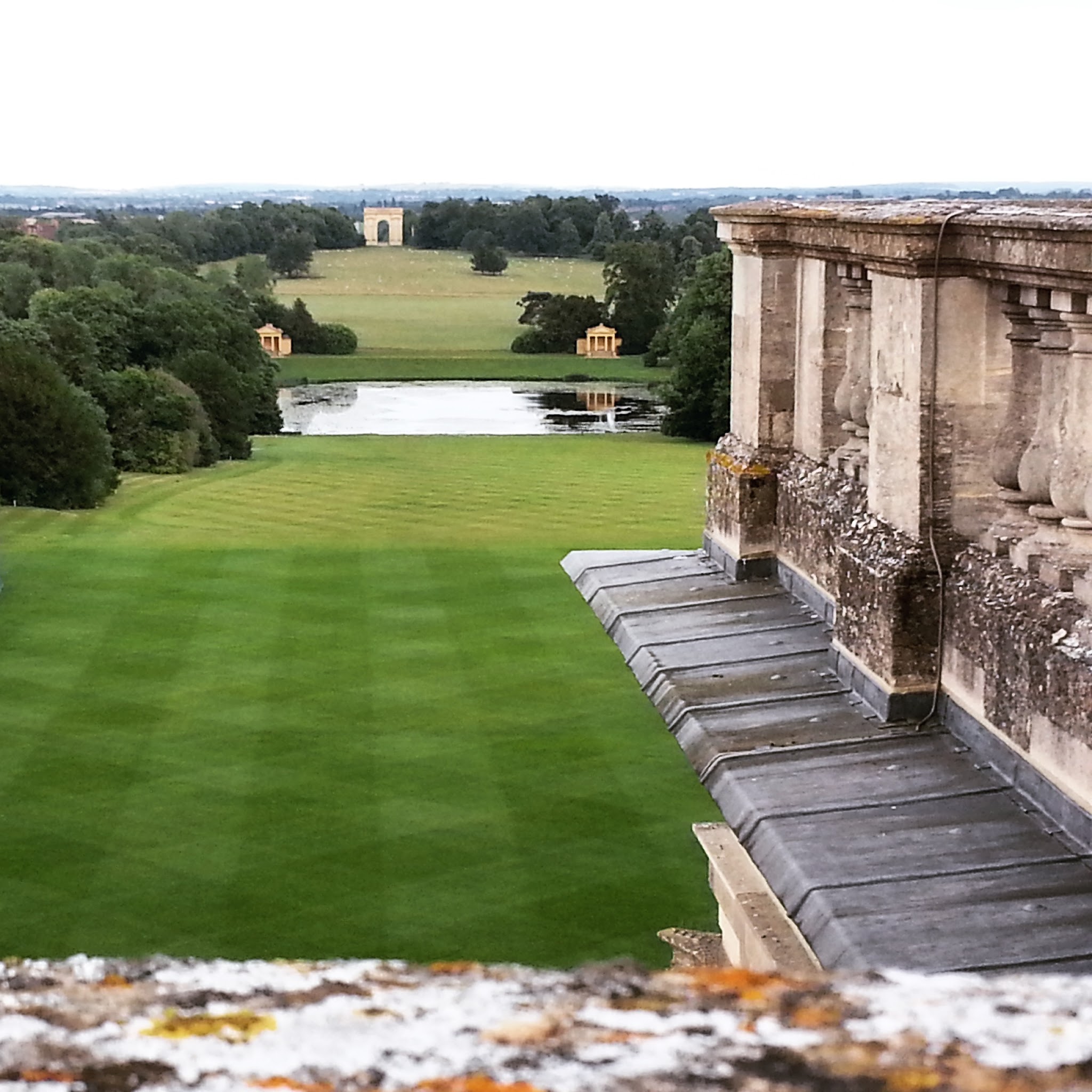 A view from high up on a roof looking across a large green lawn and towards a lake, beyond are two small temples flagging the side and beyond centre is a large golden arch monument. Trees line the views.