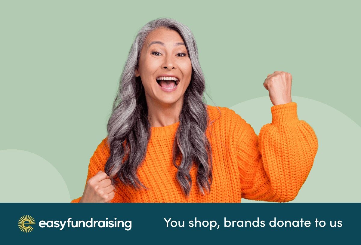 An Asian women in her late forties with long curled grey hair smiles and gestures a 'yes' closed fist like she's winning and wears a orange jumper. She's happy at raising money for charity