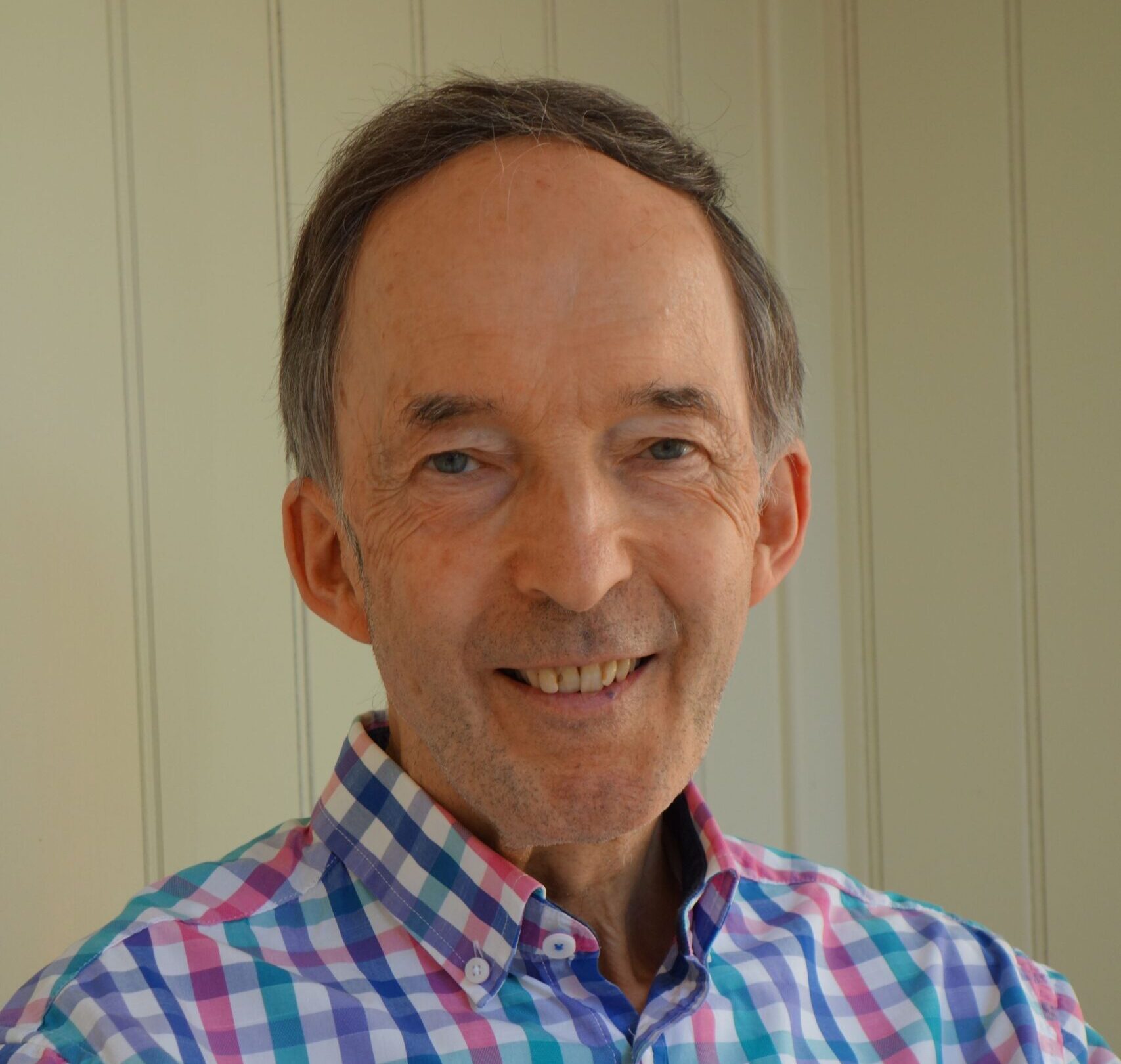 A white man in his 70s smiles, he has drak brown hair with shades of grey. He wears a bright chequered shit. He is Michael Bevington